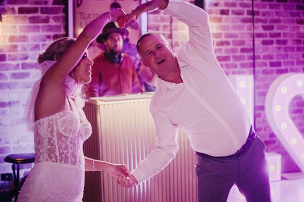 The bride Krystal dances with an older man, likely her father, at the reception stage of Sandstone Point Hotel — Cellar, with a DJ in the background and illuminated heart-shaped decor.
