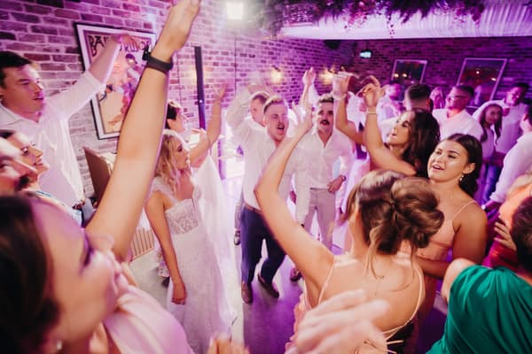 Bride Krystal in a white wedding dress and guests dance energetically at the reception stage inside Sandstone Point Hotel — Cellar with exposed brick walls and purple lighting.