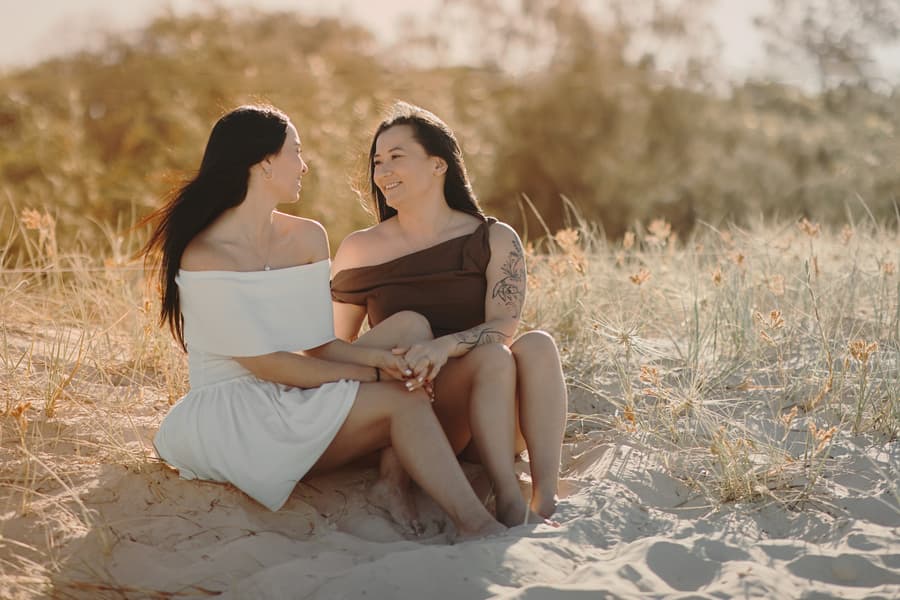 The couple sitting on sandy dunes at Woorim Beach, holding hands and smiling warmly at each other in soft sunlight.