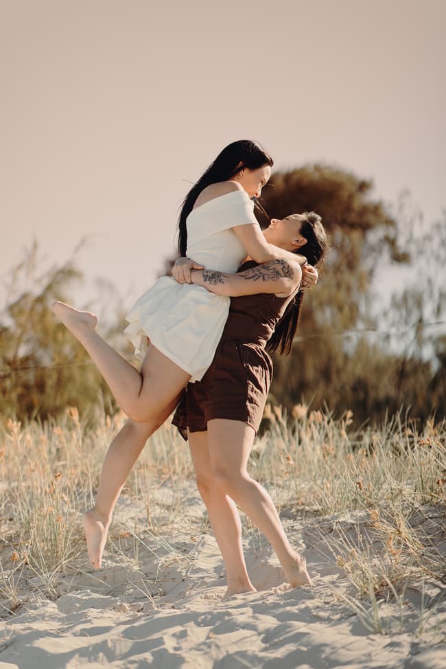 One person lifting the other playfully in the air on the sandy dunes at Woorim Beach, both laughing joyfully.