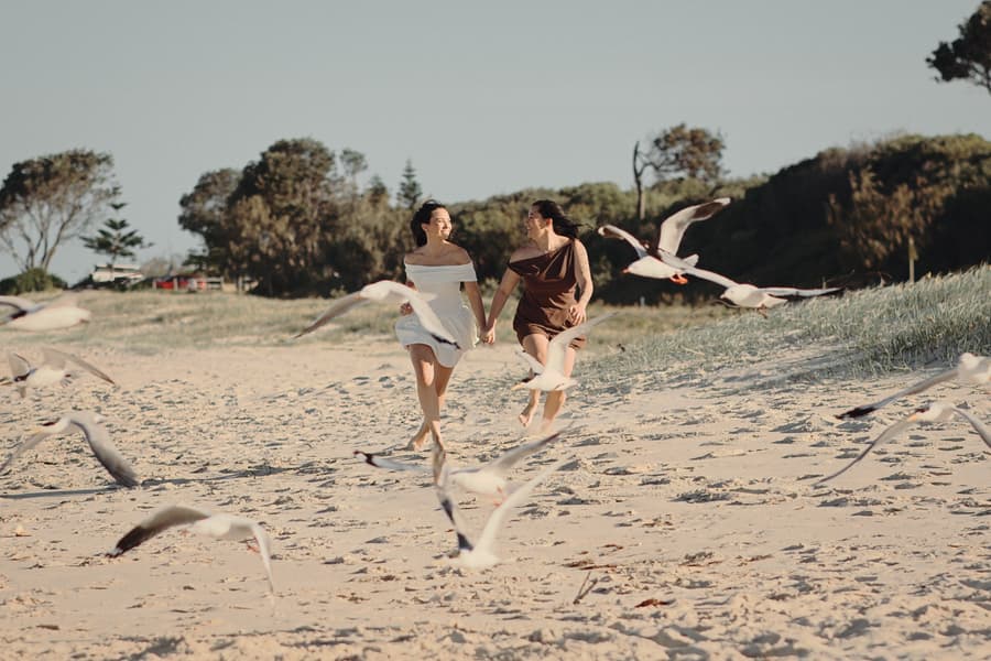 The couple running playfully through a flock of seagulls on the sandy beach at Woorim Beach, laughing together.