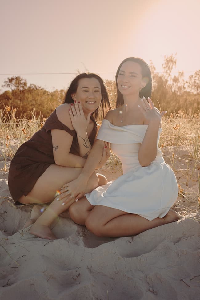 The couple kneels on the sand at Woorim Beach, smiling and showing their rings in the warm sunlight.