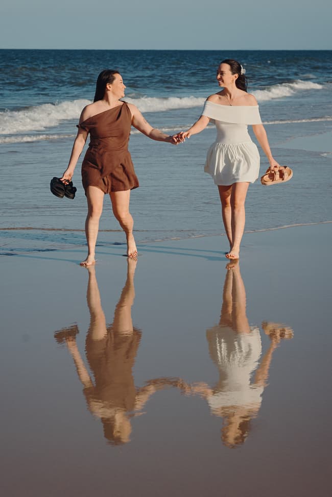 The couple holds hands while walking barefoot on the wet sand at Woorim Beach, with their reflections visible.