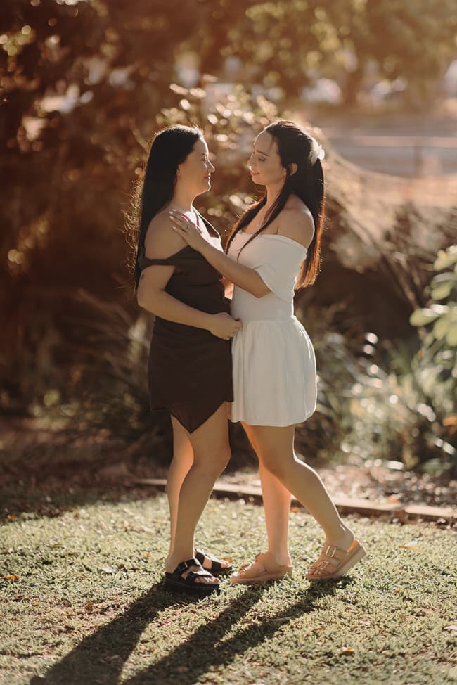 The couple stands close, gazing at each other with affection, surrounded by greenery and soft sunlight.