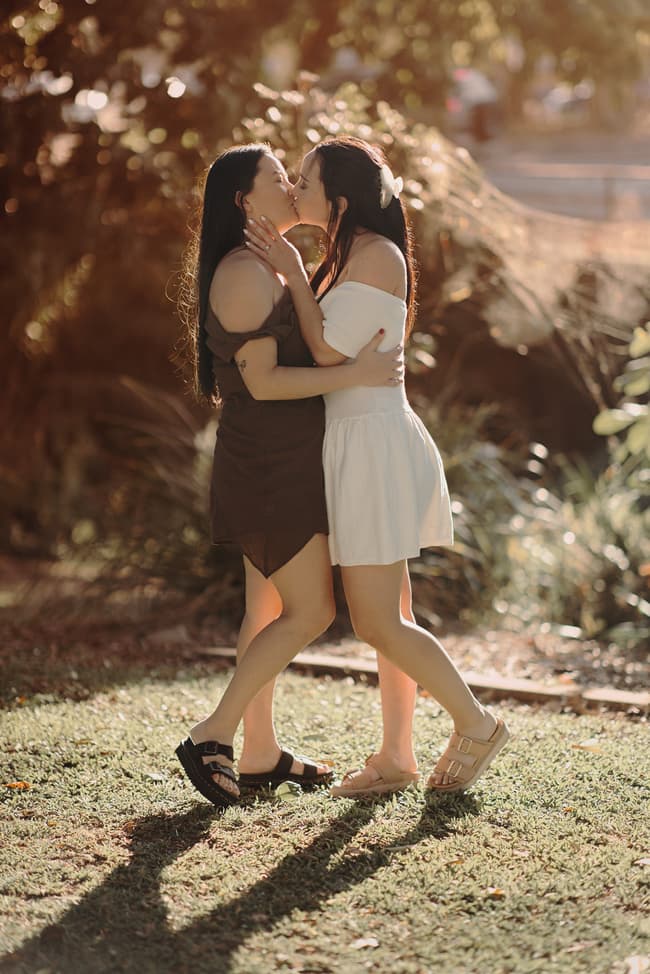 The couple shares a kiss in a sunlit garden, surrounded by lush greenery and warm light.