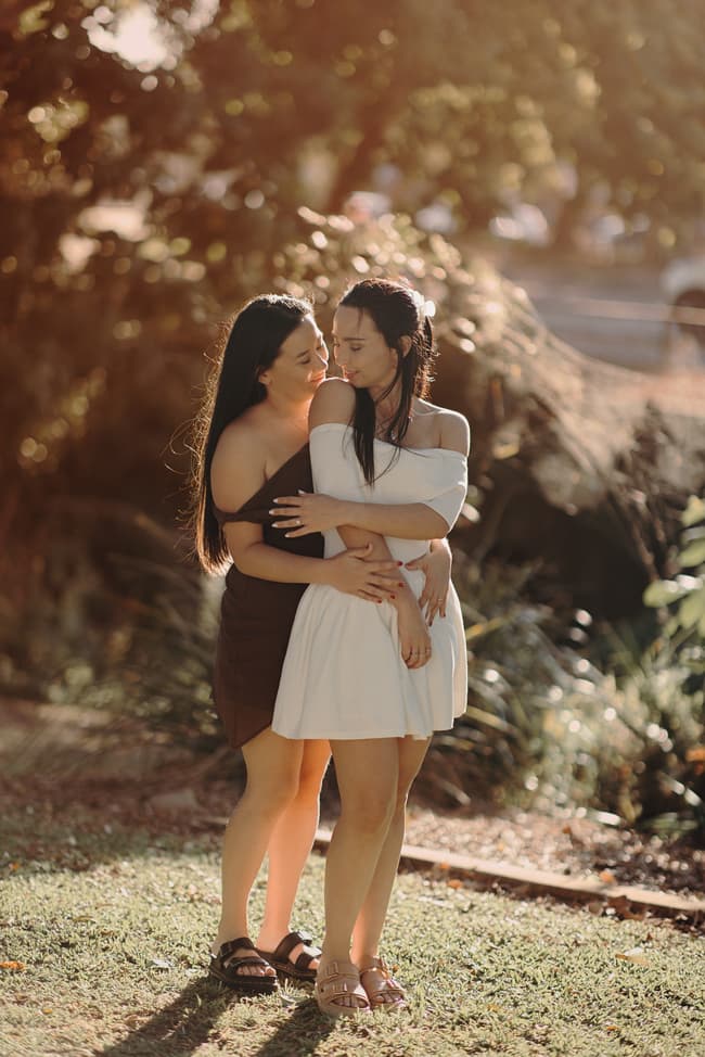 The couple embraces lovingly in a sun-dappled garden, with one resting their head on the other's shoulder.