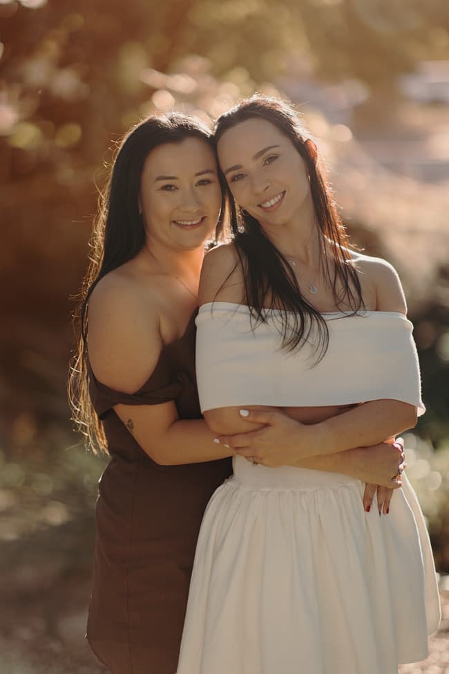 The couple stands close, smiling warmly at the camera, with sunlight filtering through the trees.