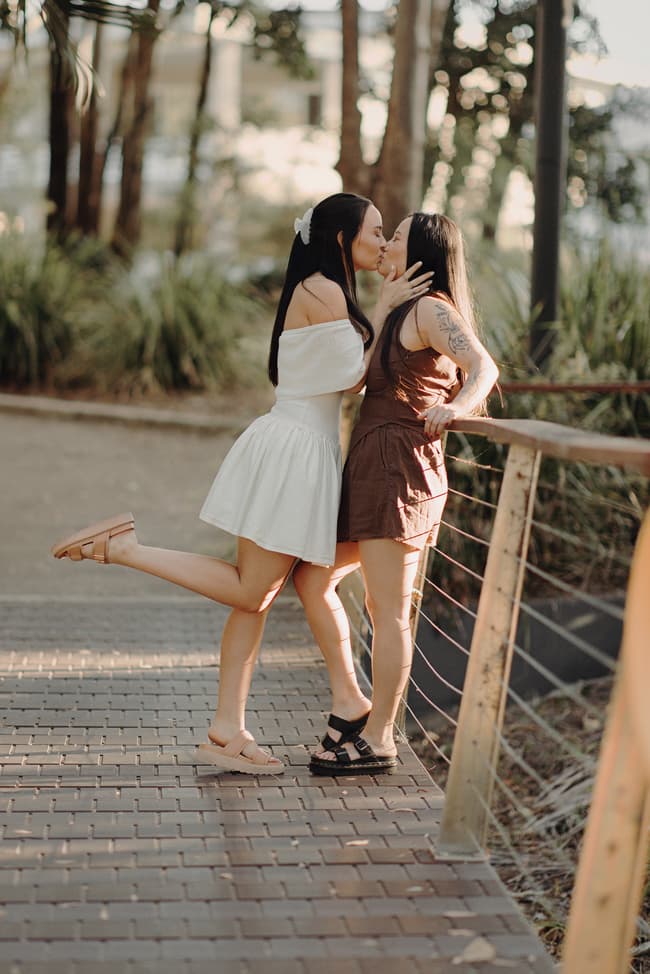 The couple shares a kiss on a wooden bridge, one lifting a foot playfully, surrounded by trees.