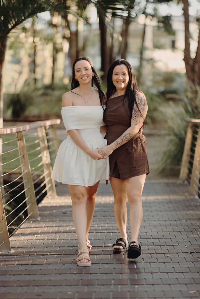 The couple stands side by side, holding hands on a wooden bridge, smiling at the camera.