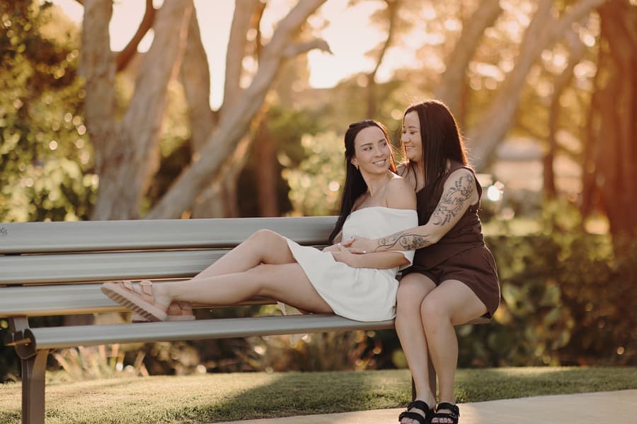 The couple sits on a park bench, one leaning back against the other, both smiling in the warm afternoon light.