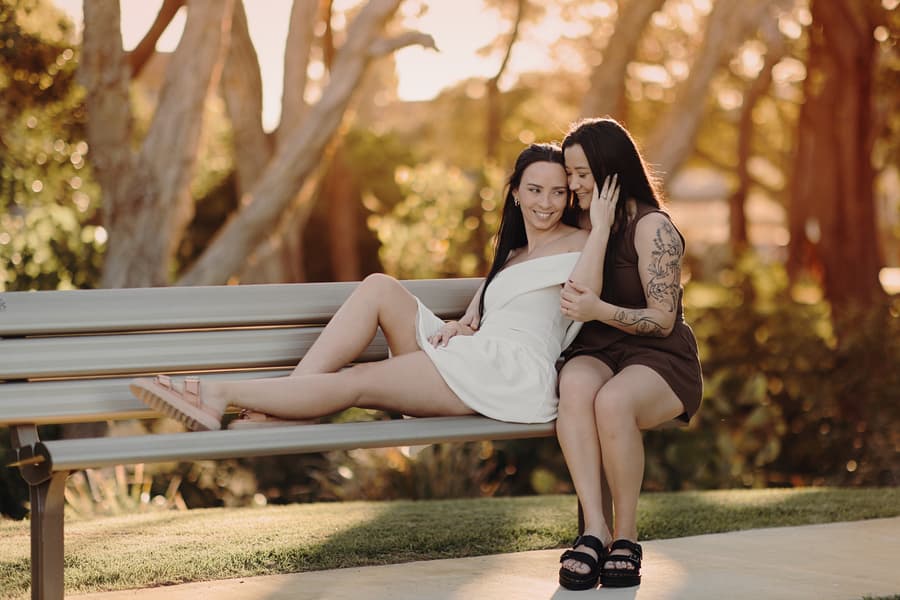 The couple sits on a park bench, one whispering to the other, both smiling under the dappled sunlight.