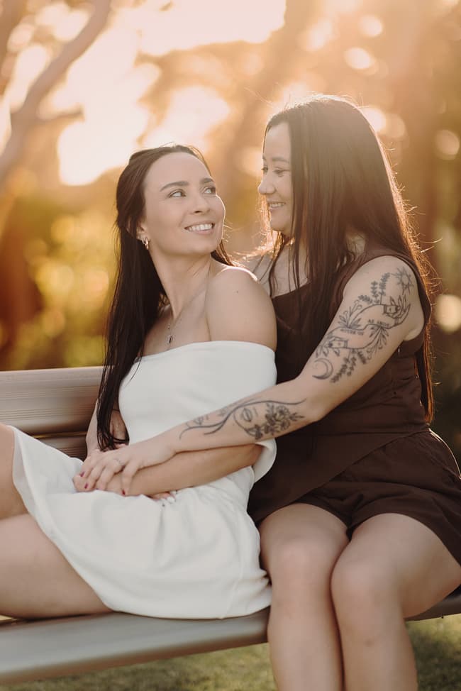 The couple sits on a bench at Woorim Beach, bathed in warm, golden sunlight. They are smiling at each other, creating a joyful and intimate atmosphere.