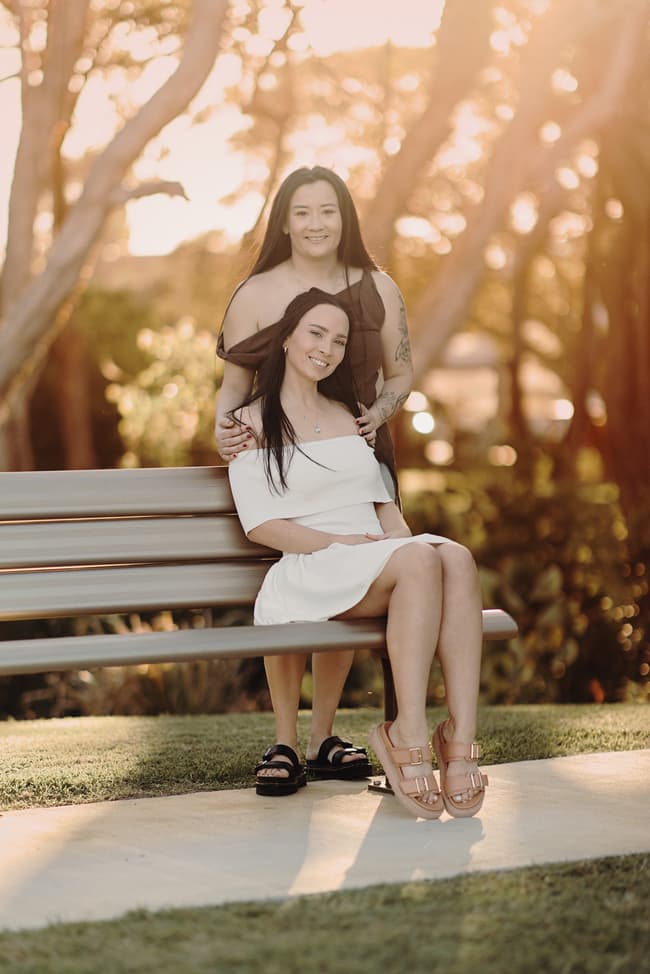 The couple poses on a bench at Woorim Beach, with one standing behind the other. The sunlight filters through the trees, adding warmth to the scene.