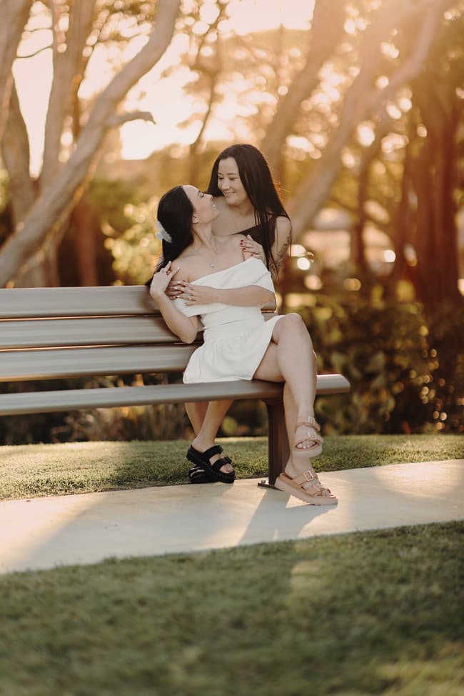 The couple sits closely on a bench at Woorim Beach, sharing a tender embrace. The warm sunlight creates a romantic ambiance.
