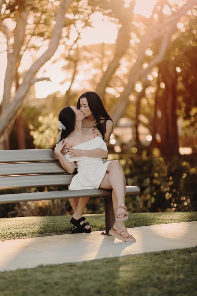 The couple shares a kiss on a bench at Woorim Beach, surrounded by soft, golden light. The scene is intimate and loving.