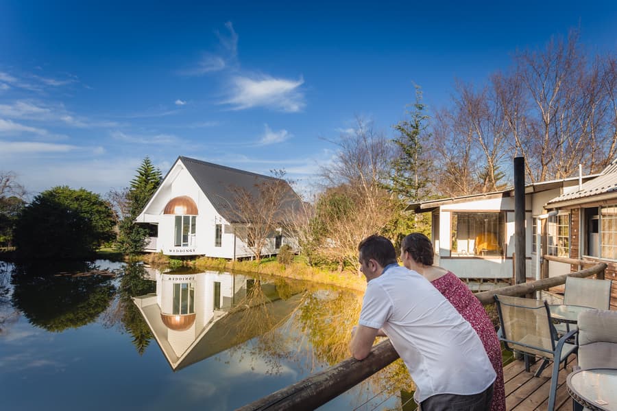 The couple leaning on a wooden railing, gazing at the reflection of a white chapel on the lake at The Abbey Cobaki.