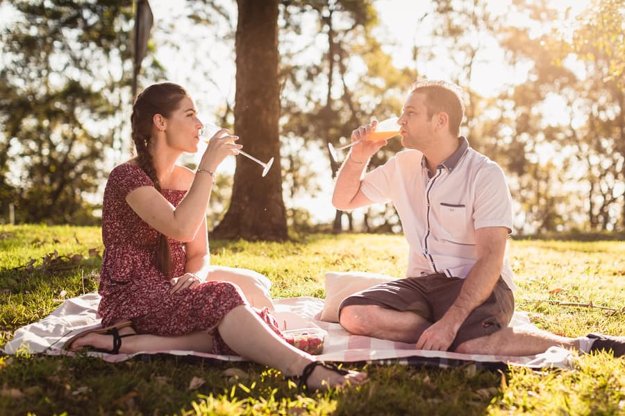 The couple sitting on a picnic blanket, toasting with drinks under the trees at The Abbey Cobaki.