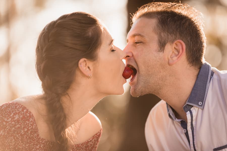 The couple playfully share a strawberry with their mouths in warm, golden light at The Abbey Cobaki.
