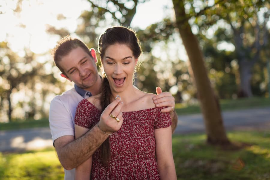 The couple stands in a park, one showing surprise as the other presents a ring, with sunlight filtering through trees.