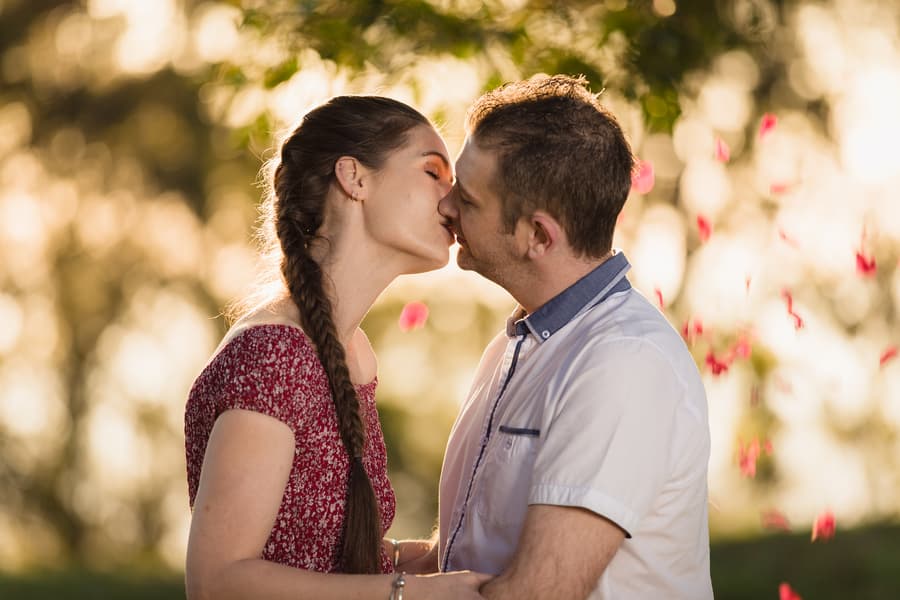 The couple shares a kiss surrounded by falling petals, with a sunlit background at The Abbey Cobaki.