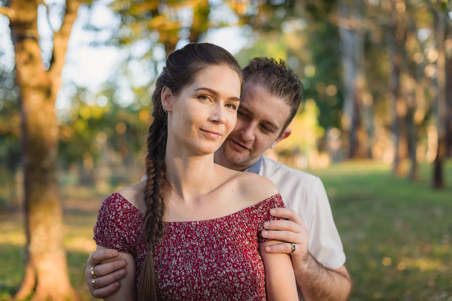 The couple stands close together, one resting their head on the other's shoulder, in a sunlit park.