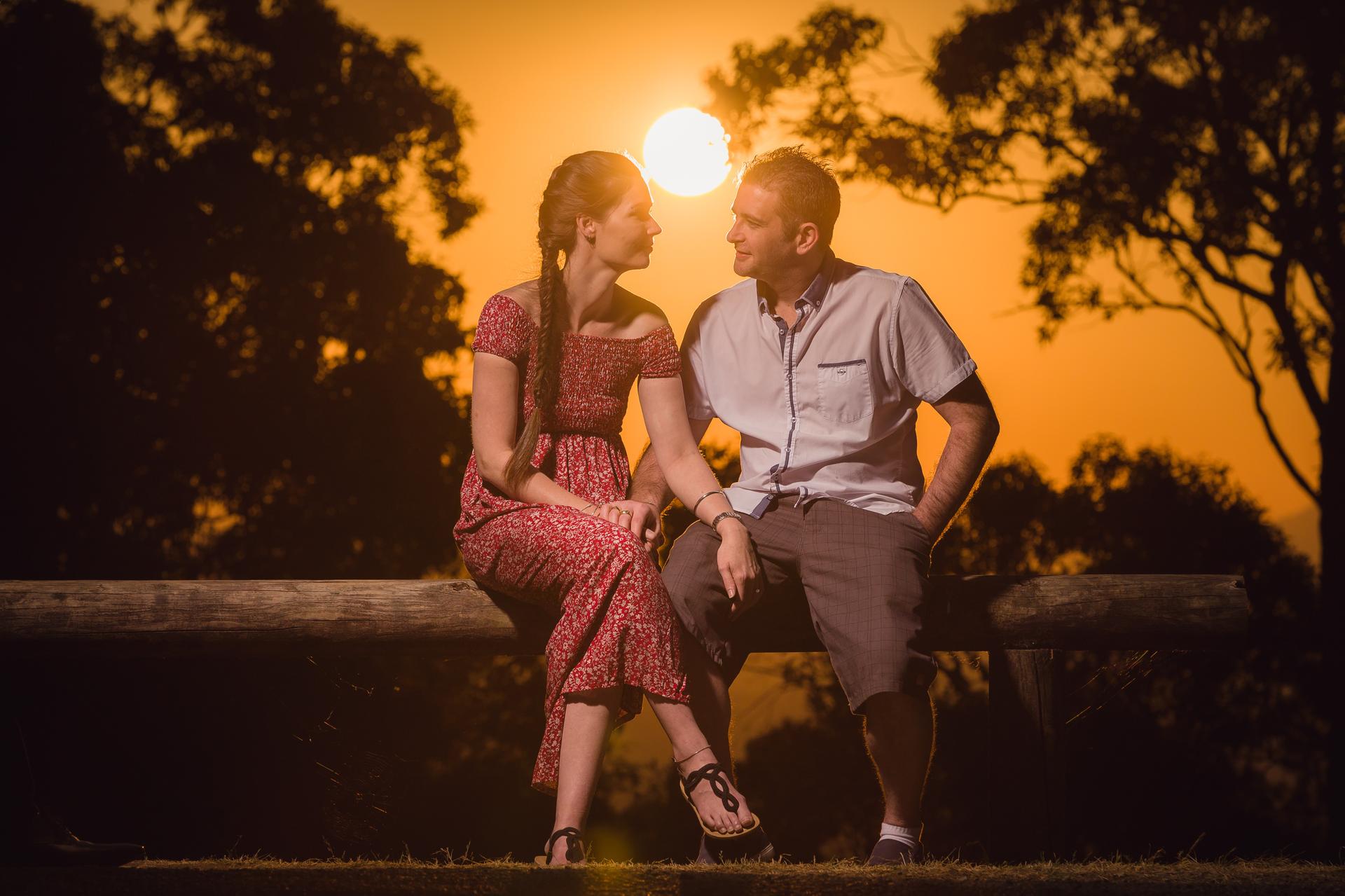 The couple sits on a wooden bench, silhouetted against a vibrant sunset, gazing at each other.
