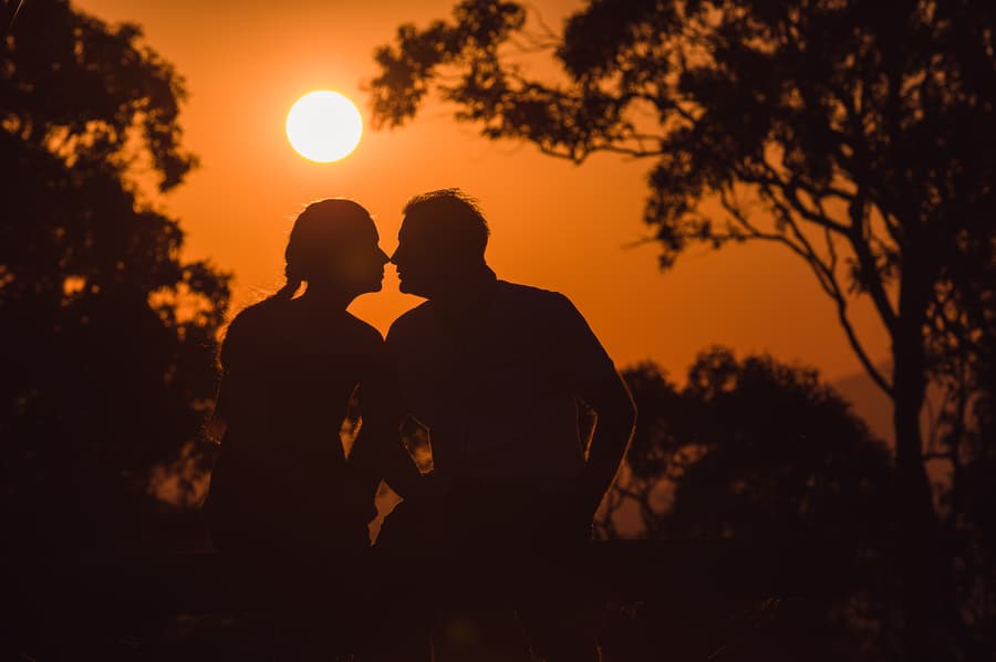 The couple shares a silhouette kiss against a vivid orange sunset, with trees framing the scene.