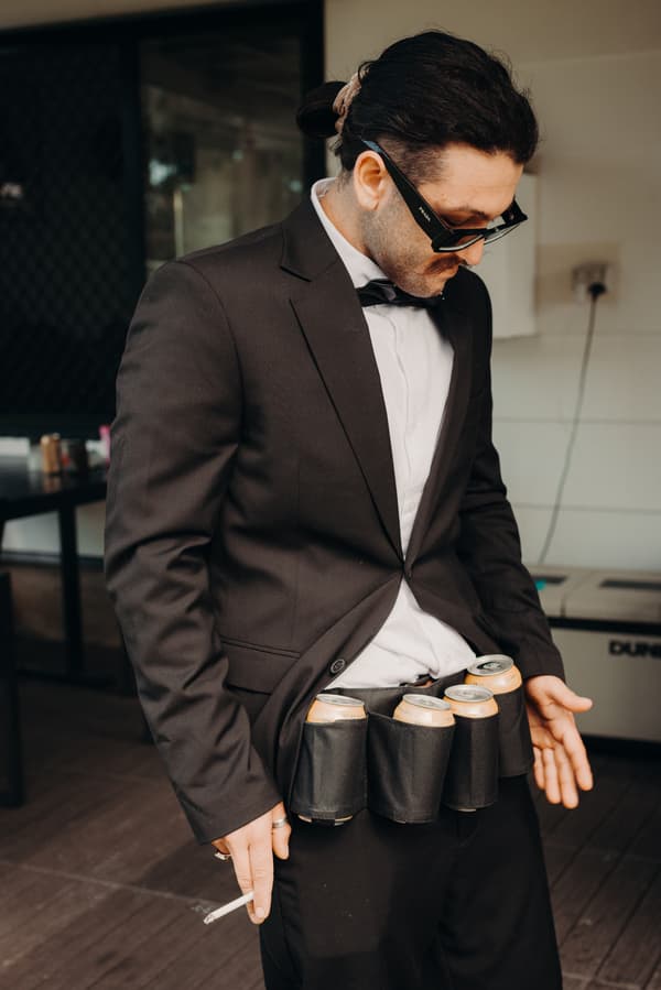 The groom Kyle stands indoors wearing a black suit and sunglasses, with a belt holding four cans around his waist and a cigarette in his left hand.