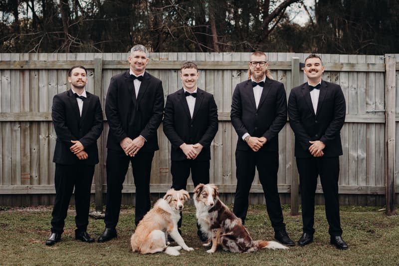 Five groomsmen stand in a row outdoors in front of a wooden fence at The Tides, wearing black tuxedos with bow ties, with two dogs sitting on the grass in front of them.