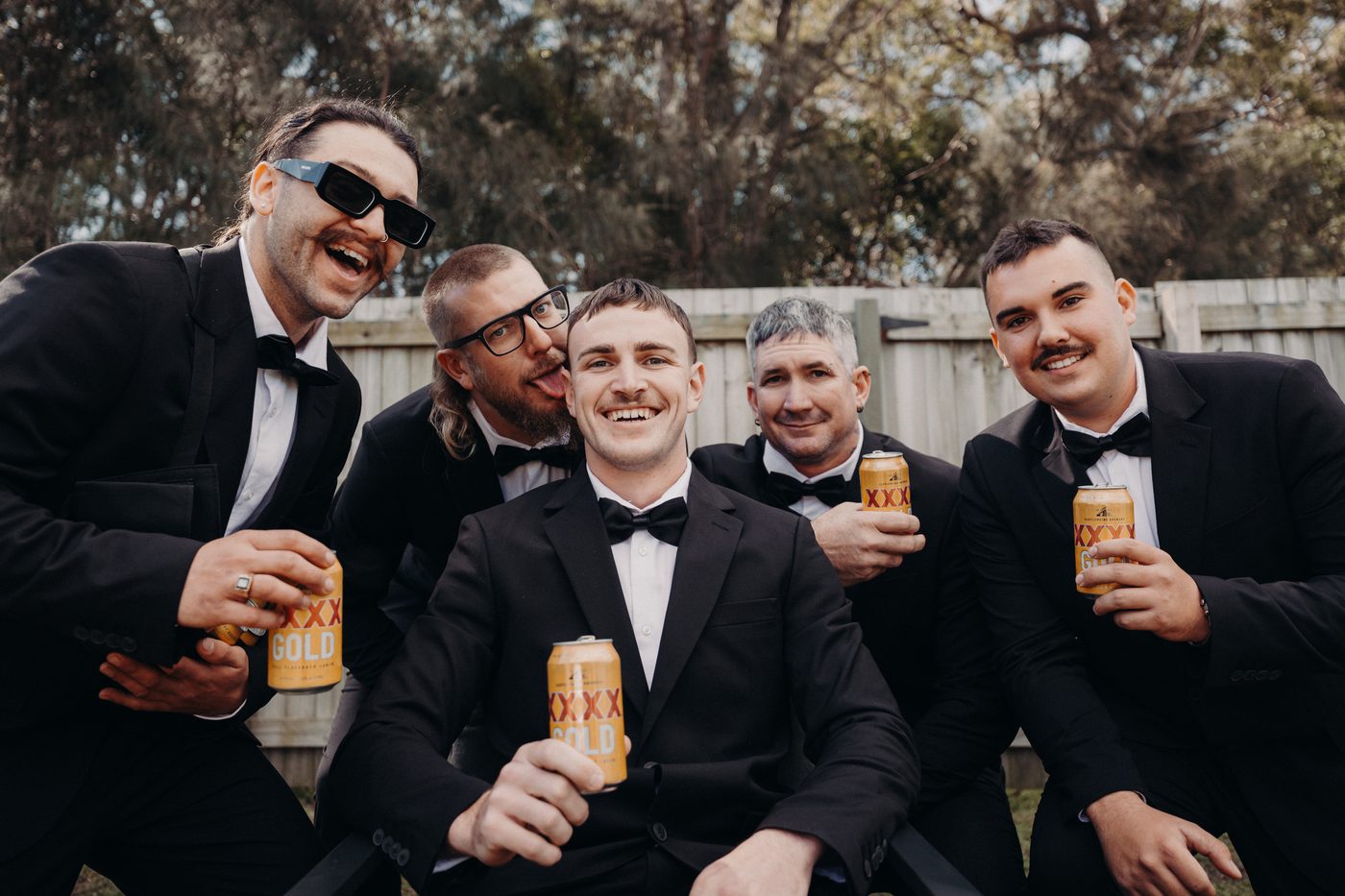 Kyle and four groomsmen dressed in black tuxedos pose outdoors holding cans of XXXX Gold beer in front of a wooden fence with trees in the background. Wedding fair season Brisbane by She Said Yes.