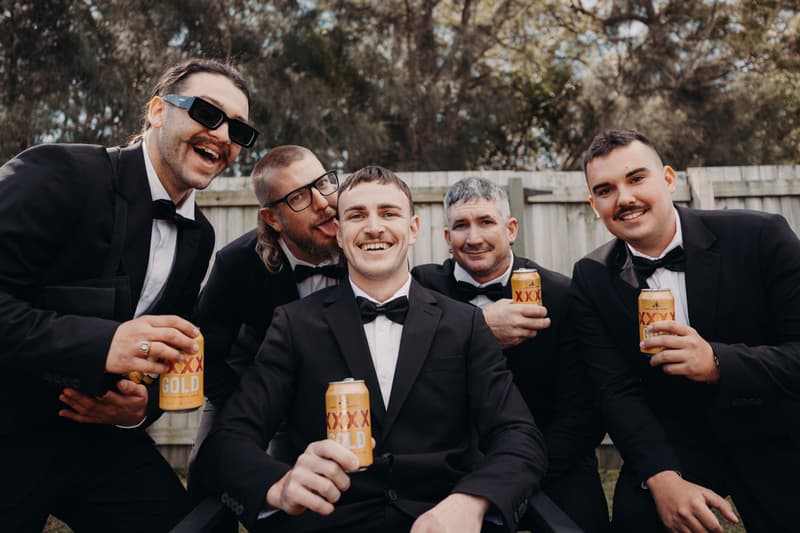 Kyle and four groomsmen dressed in black tuxedos pose outdoors holding cans of XXXX Gold beer in front of a wooden fence with trees in the background.