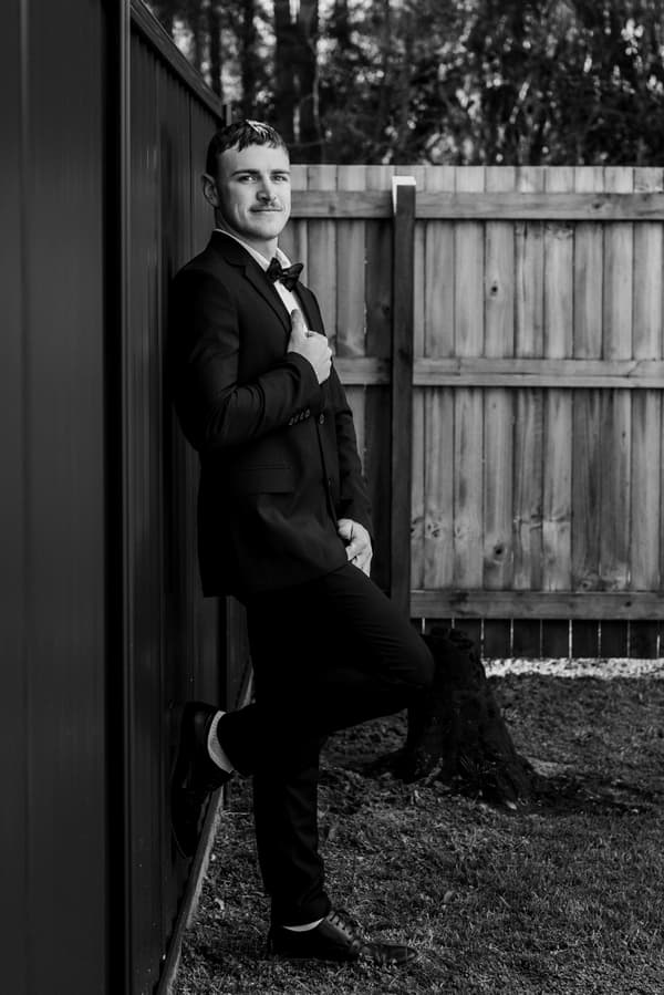 The groom, Kyle, leans against a dark fence outdoors near a wooden fence, dressed in a suit with a bow tie.