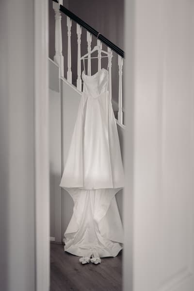The bride's wedding dress hangs on a staircase banister with matching white shoes placed on the floor beneath it. Wedding live streaming by She Said Yes.