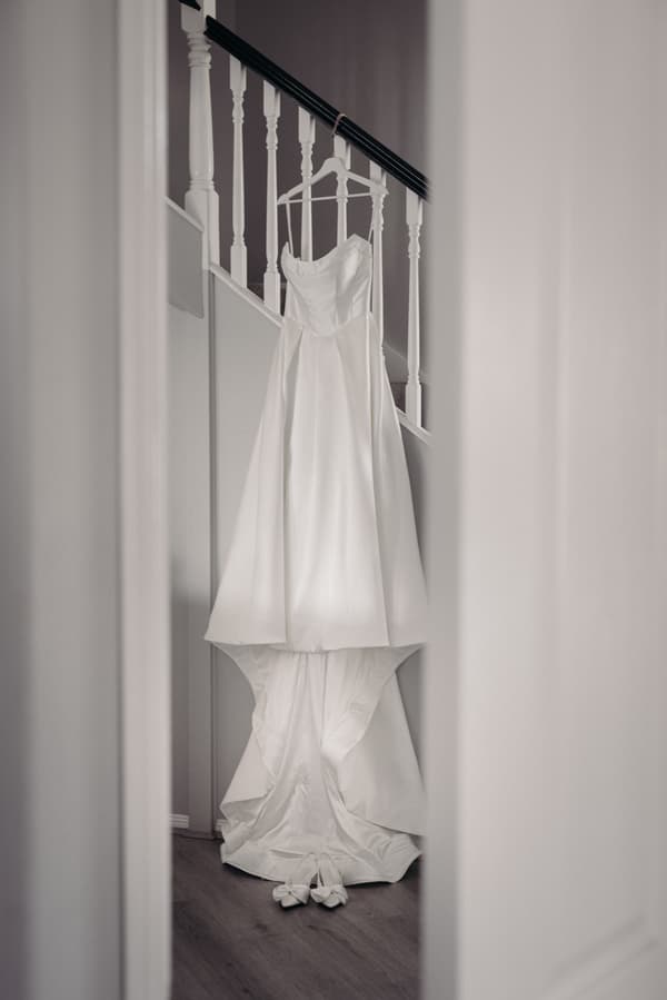 The bride's wedding dress hangs on a staircase banister with matching white shoes placed on the floor beneath it.