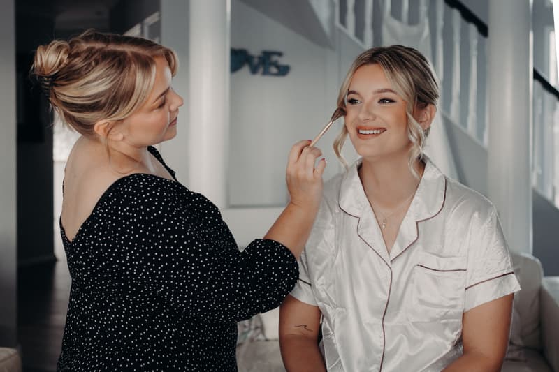 The bride Libby sits while a woman applies makeup to her face indoors near a staircase.