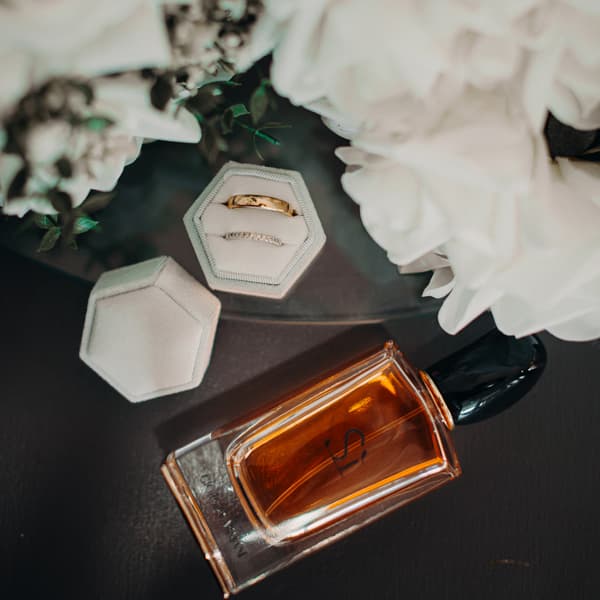 Wedding rings in a hexagonal white ring box next to a bottle of amber-colored perfume and white flowers.