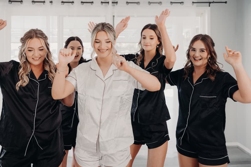 The bride Libby in white pajamas dances with four bridesmaids in matching black pajamas in a bright room at The Tides.