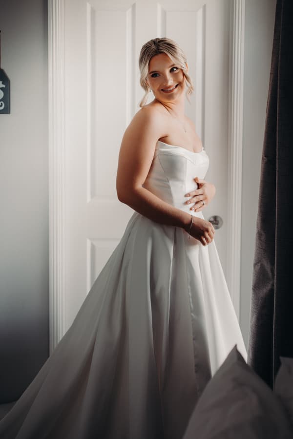 The bride Libby poses in her wedding gown in front of a white door indoors.