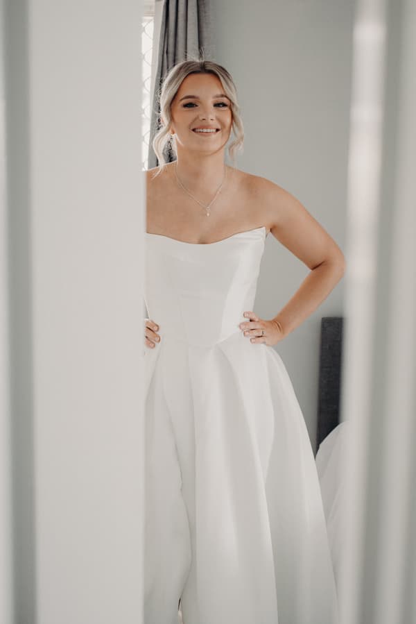 The bride Libby stands indoors wearing a strapless white wedding gown with her hands on her hips, smiling.