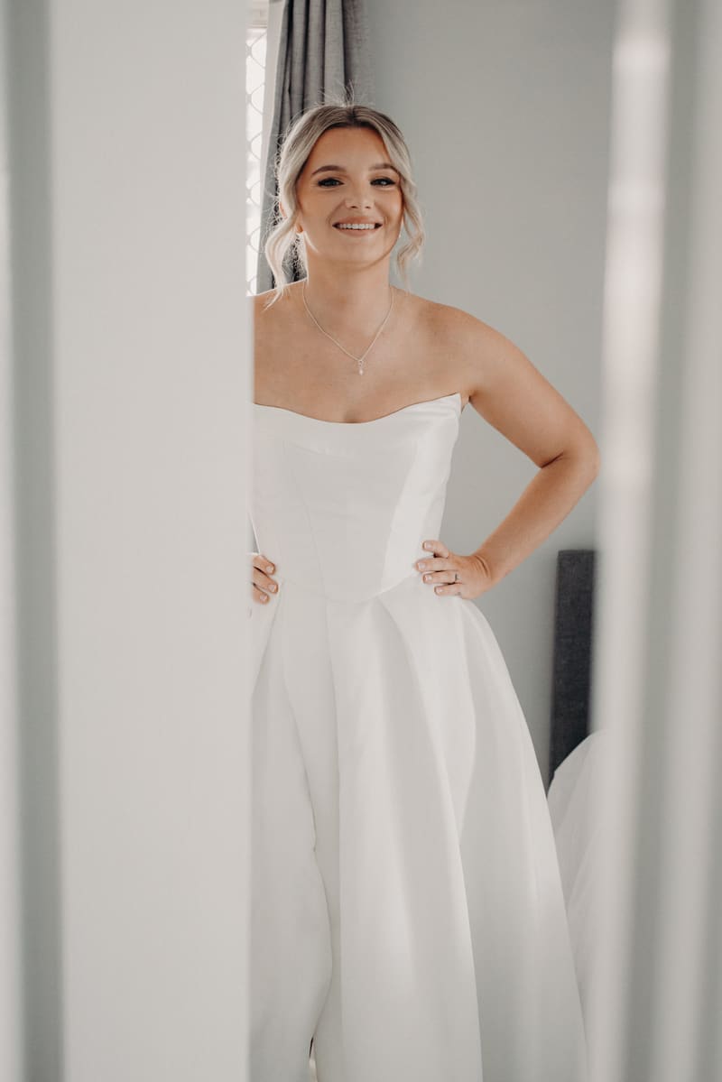 The bride Libby stands indoors wearing a strapless white wedding gown with her hands on her hips, smiling. Wedding photography contracts by She Said Yes.