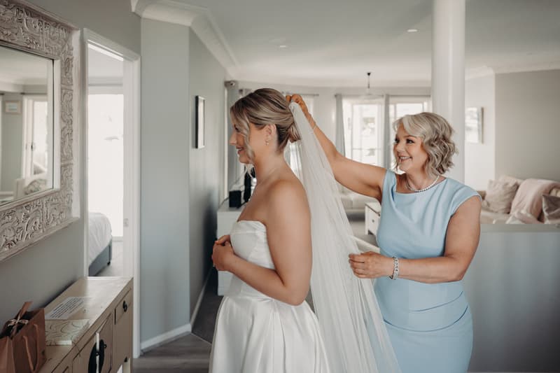 The bride Libby stands in a room at The Tides preparing for the wedding while an older woman, likely the mother of the bride, adjusts her veil.