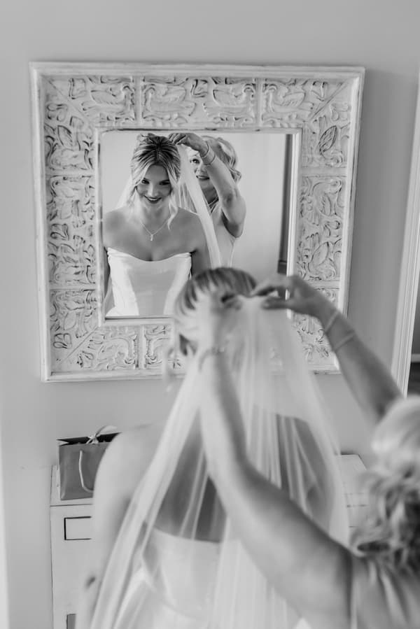 The bride Libby is having her veil adjusted by an older woman, possibly her mother, reflected in a decorative mirror.