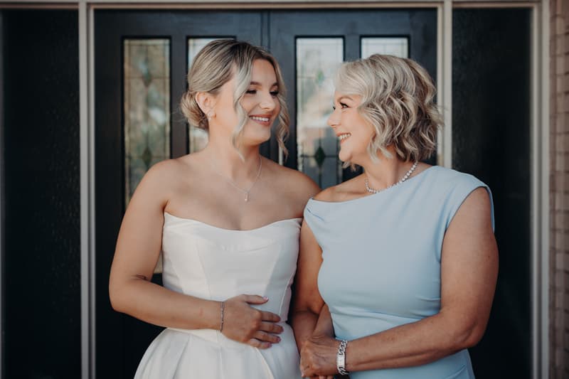 The bride Libby stands with the mother of the bride in front of a dark door with decorative glass panels at The Tides — The Water's Edge.