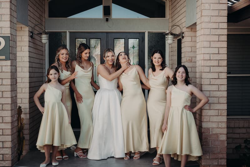 Libby the bride poses with six bridesmaids in cream dresses outside The Tides — The Water's Edge entrance.