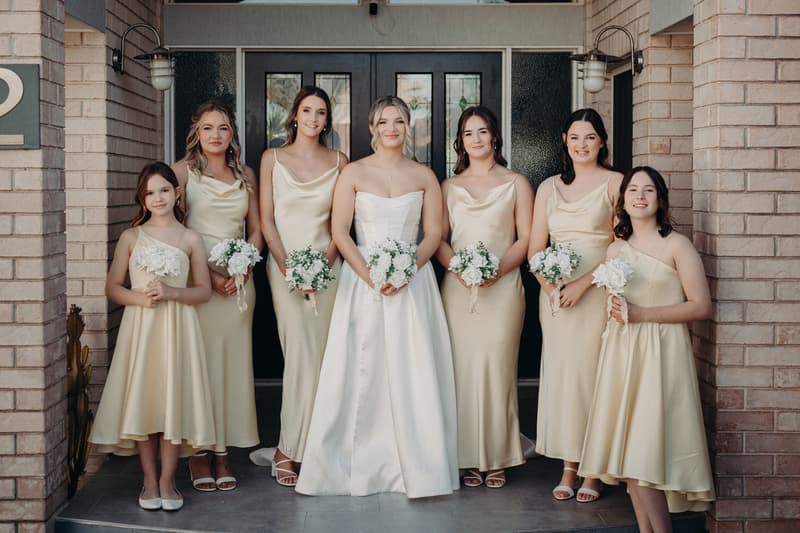 The bride Libby stands with six bridesmaids and two flower girls in cream-colored dresses holding bouquets outside a building with brick walls and double glass doors at The Tides — The Water's Edge.