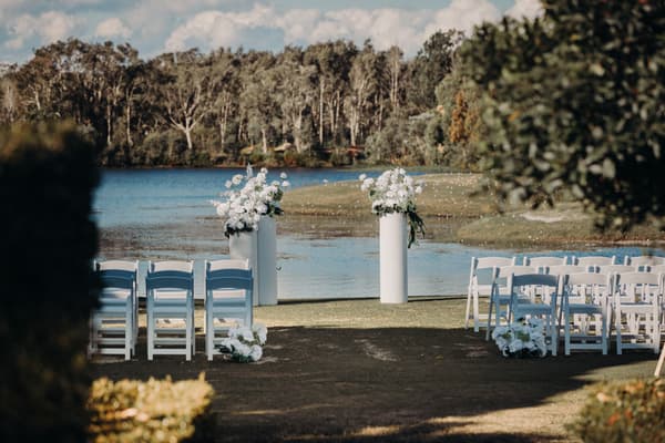 Empty outdoor wedding ceremony setup at The Tides — The Water's Edge with white chairs arranged in rows facing two white floral arrangements on tall stands by the water.