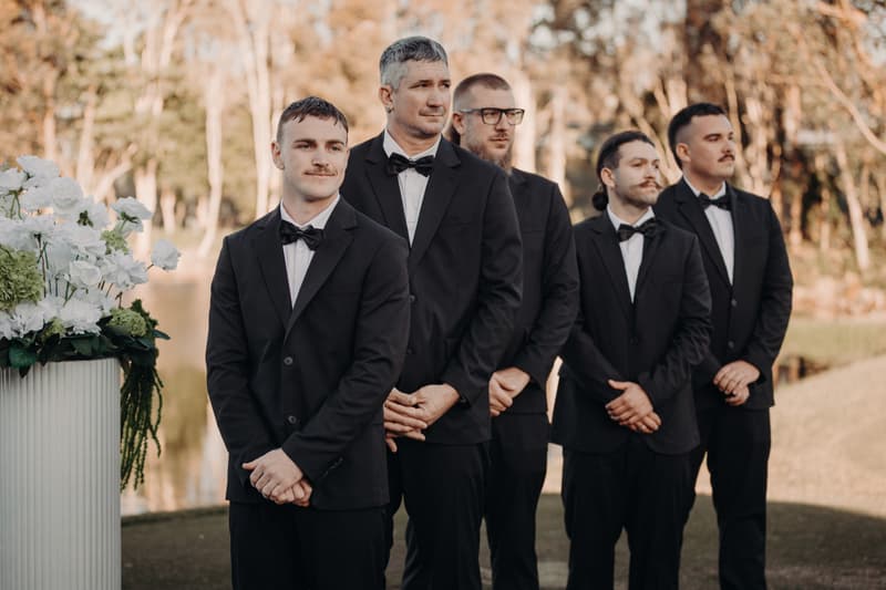 Five groomsmen stand in a line outdoors at The Tides — The Water's Edge, dressed in black tuxedos with bow ties, near a floral arrangement with white flowers and greenery.
