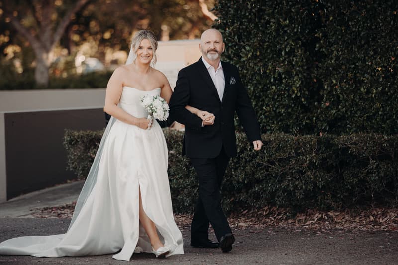 The bride Libby walks arm-in-arm with an older man, likely her father, along an outdoor path at The Tides — The Water's Edge. Libby wears a strapless white wedding gown and holds a small bouquet of white flowers, while the man wears a black suit with a white shirt.