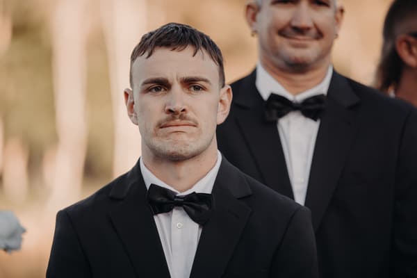 Kyle, the groom, stands in a black tuxedo with a bow tie at The Tides — The Water's Edge during the ceremony stage, with groomsmen partially visible behind him.