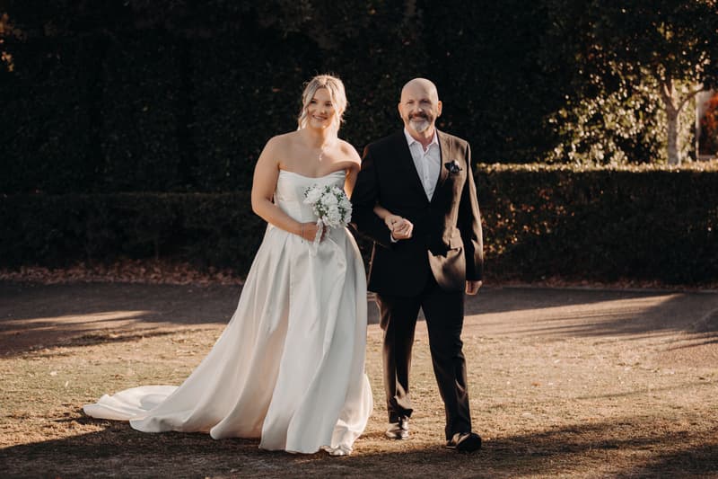 The bride Libby walks arm-in-arm with an older man, likely her father, outdoors at The Tides — The Water's Edge, holding a bouquet of white flowers.